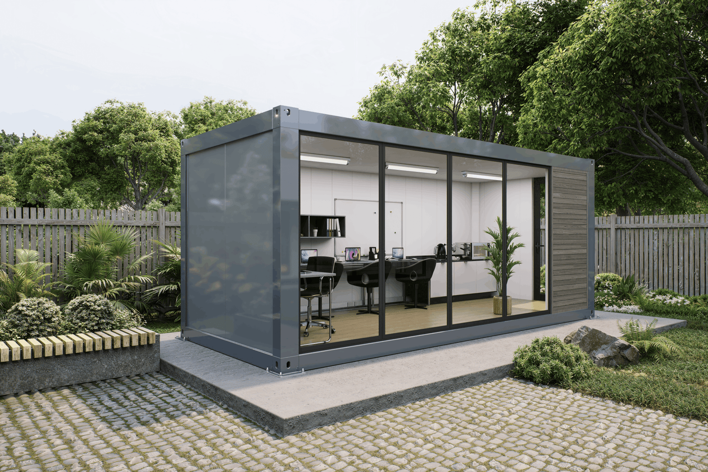 A modern modular office pod made from a repurposed shipping container, featuring a dark gray metal frame, large glass windows, and a wood-paneled exterior. The pod is situated on a raised concrete platform in a landscaped garden with a stone pathway, green plants, and a wooden fence in the background.