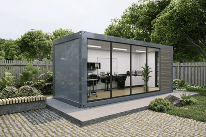 A modern modular office pod made from a repurposed shipping container, featuring a dark gray metal frame, large glass windows, and a wood-paneled exterior. The pod is situated on a raised concrete platform in a landscaped garden with a stone pathway, green plants, and a wooden fence in the background.
