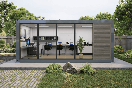 A modern modular office pod with large glass windows and a dark gray metal frame, set on a concrete patio in a landscaped garden with trees and grass.
