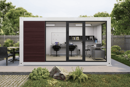 A modern modular office pod with a white exterior, dark brown wood paneling, and black-framed sliding glass doors, shown in a landscaped backyard setting with a furnished office interior visible inside.