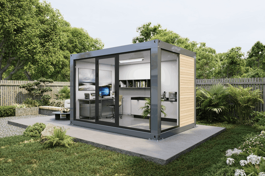A modern modular office pod with a dark gray metal frame, glass walls, and light wood siding, situated on a concrete pad in a landscaped garden setting.