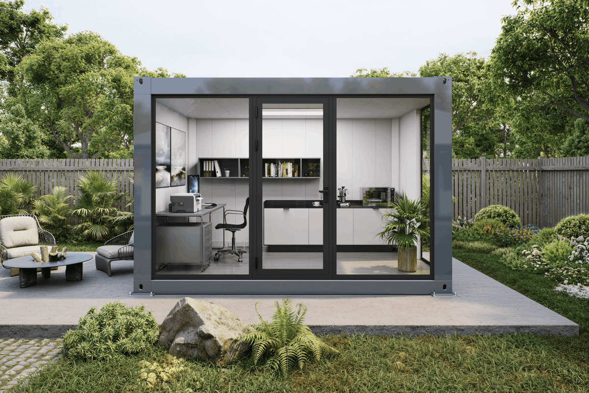 A modern modular office pod with a dark gray metal frame and large glass doors, set on a concrete patio in a landscaped garden. The office interior features white cabinets, a desk, shelving, and a kitchenette.