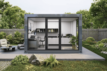A modern modular office pod with a dark gray metal frame and large glass doors, set on a concrete patio in a landscaped garden. The office interior features white cabinets, a desk, shelving, and a kitchenette.